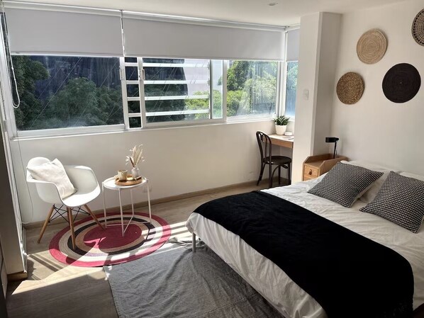 Ein Schlafzimmer mit Bett, Sessel, kleinem Tisch, Fenster mit Blick auf Grünflächen und Wandgestaltung.