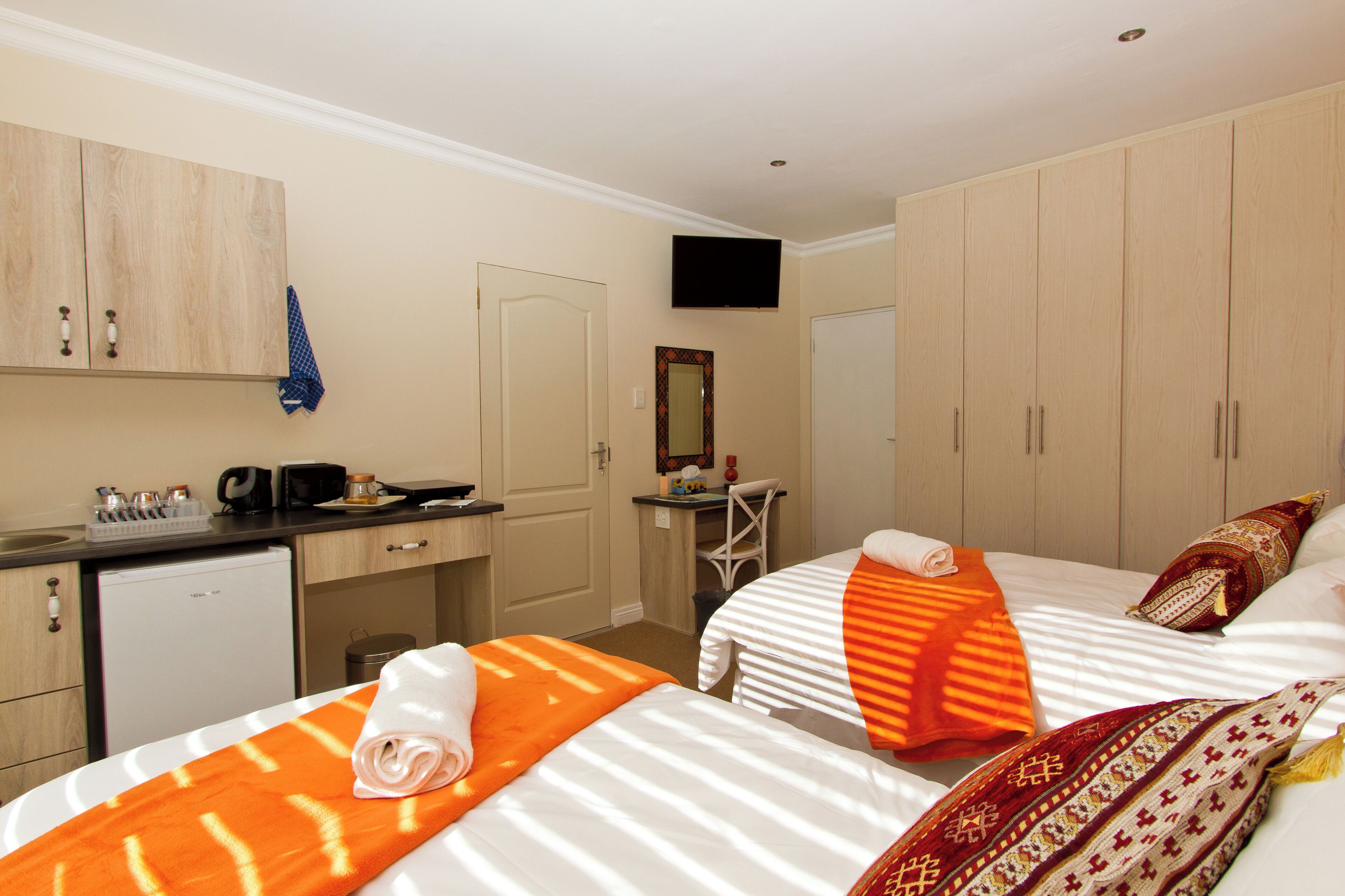 A hotel room with two beds, a desk, a small kitchenette, and a TV mounted on the wall.
