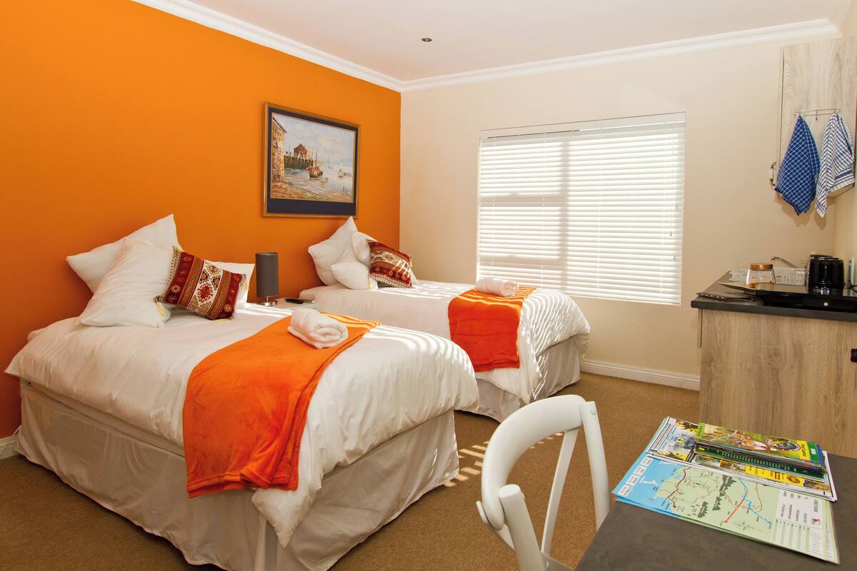 A hotel room with two beds, an orange wall, a painting, a window with blinds, a desk with a map, and a chair.