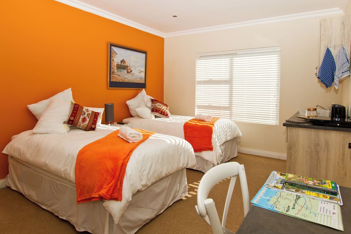 A hotel room with two beds, an orange wall, a painting, a window with blinds, a desk with a map, and a chair.