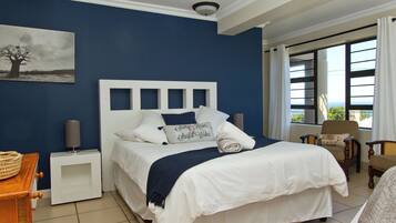 Un dormitorio con una cama grande, una mesita de noche, una silla, una ventana con cortinas y un cuadro en la pared.