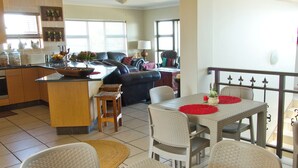 A dining area with wicker furniture, a kitchen in the background, and a balcony with a railing.