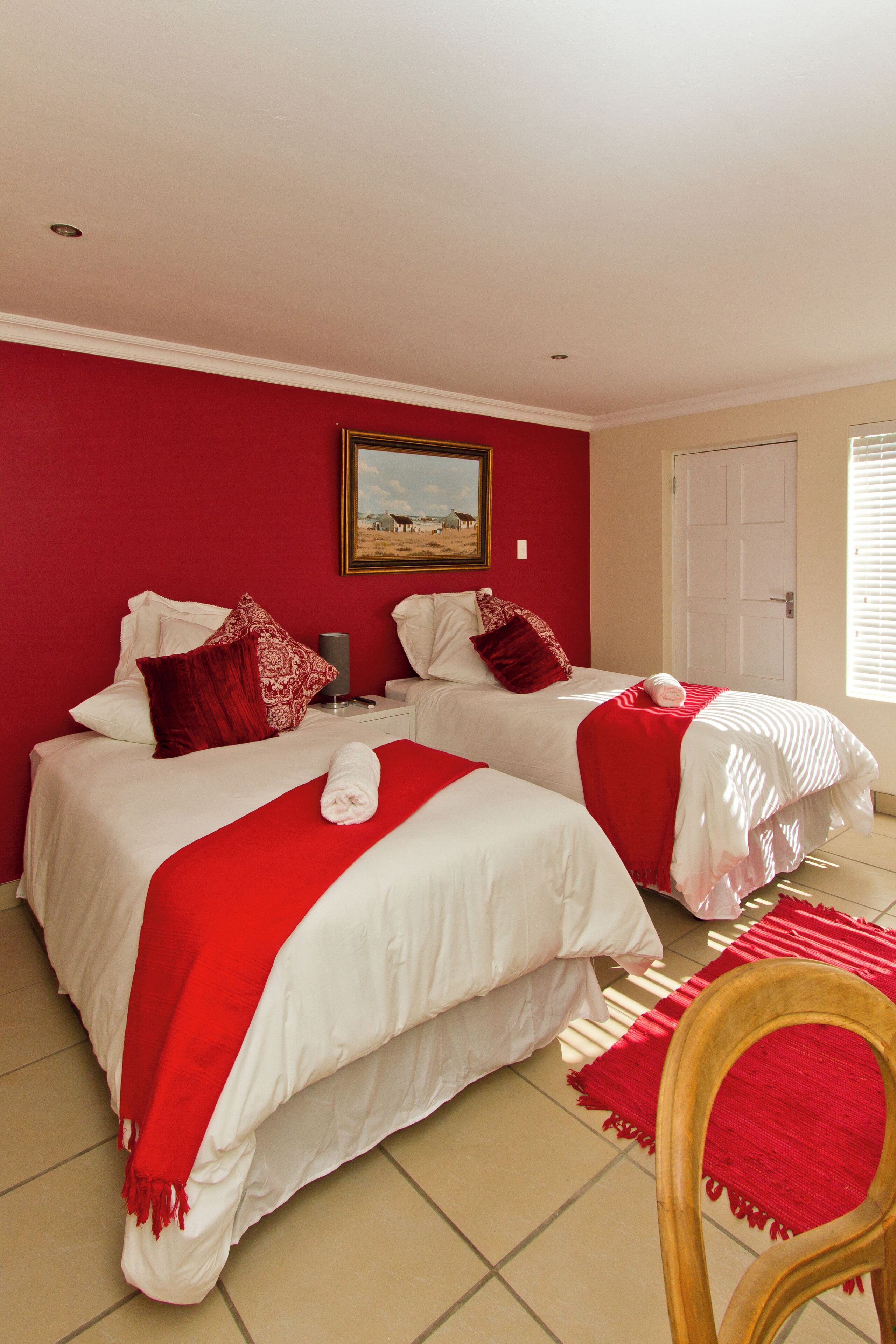 A hotel room with two beds, red and white bedding, a wooden chair, and a painting on the wall.