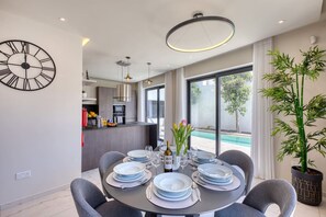 Ein moderner Essbereich mit einem für sechs Personen gedeckten Rundtisch, eine Küche im Hintergrund und ein großes Fenster mit Blick auf einen Swimmingpool.