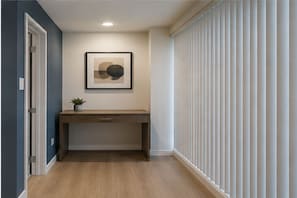A hallway with a desk, a framed artwork, a potted plant, and a window with blinds. - Cendelle City Suites (Toronto)