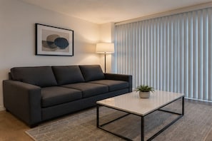 A modern living room with a dark gray sofa, a white coffee table, a floor lamp, a framed abstract artwork, and a window with vertical blinds. - Cendelle City Suites (Toronto)