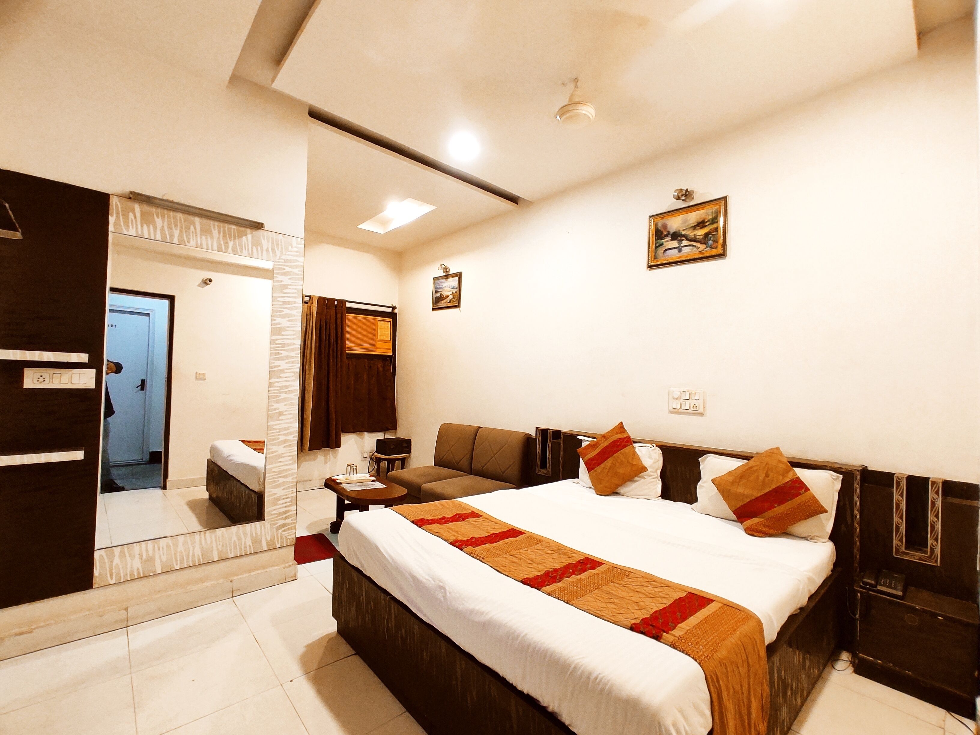 Habitación de hotel con cama, mesita de noche, espejo, ventana con cortinas y un cuadro en la pared.