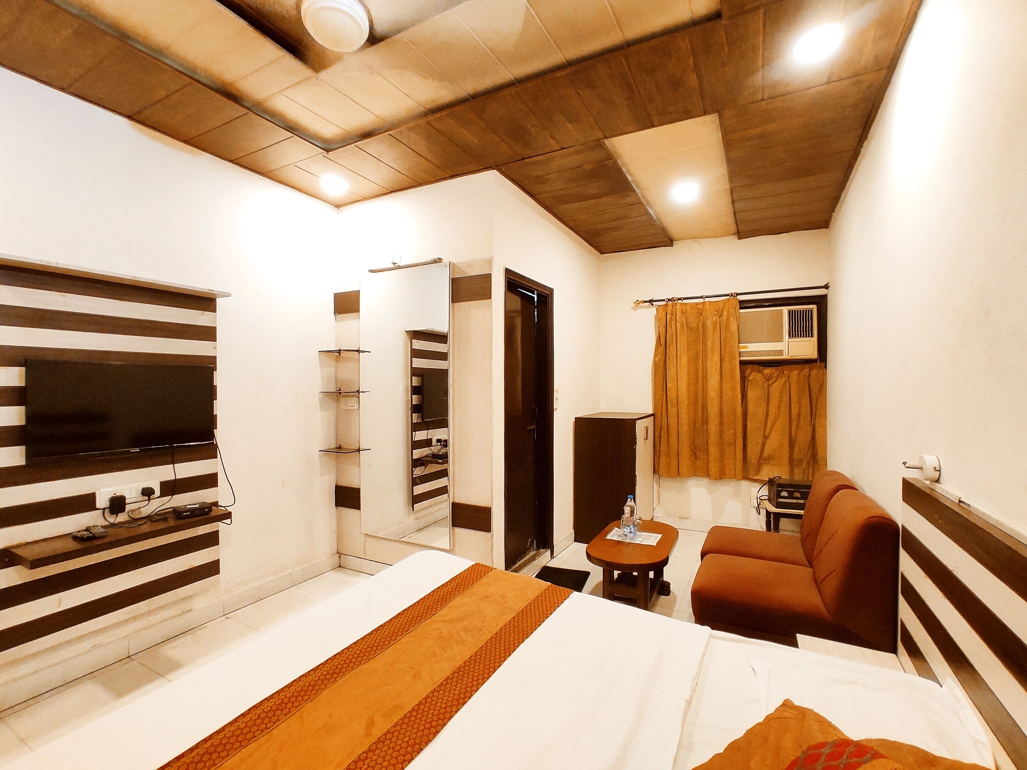 A hotel room with a bed, a TV mounted on the wall, a sofa, a small table, and a mirror.