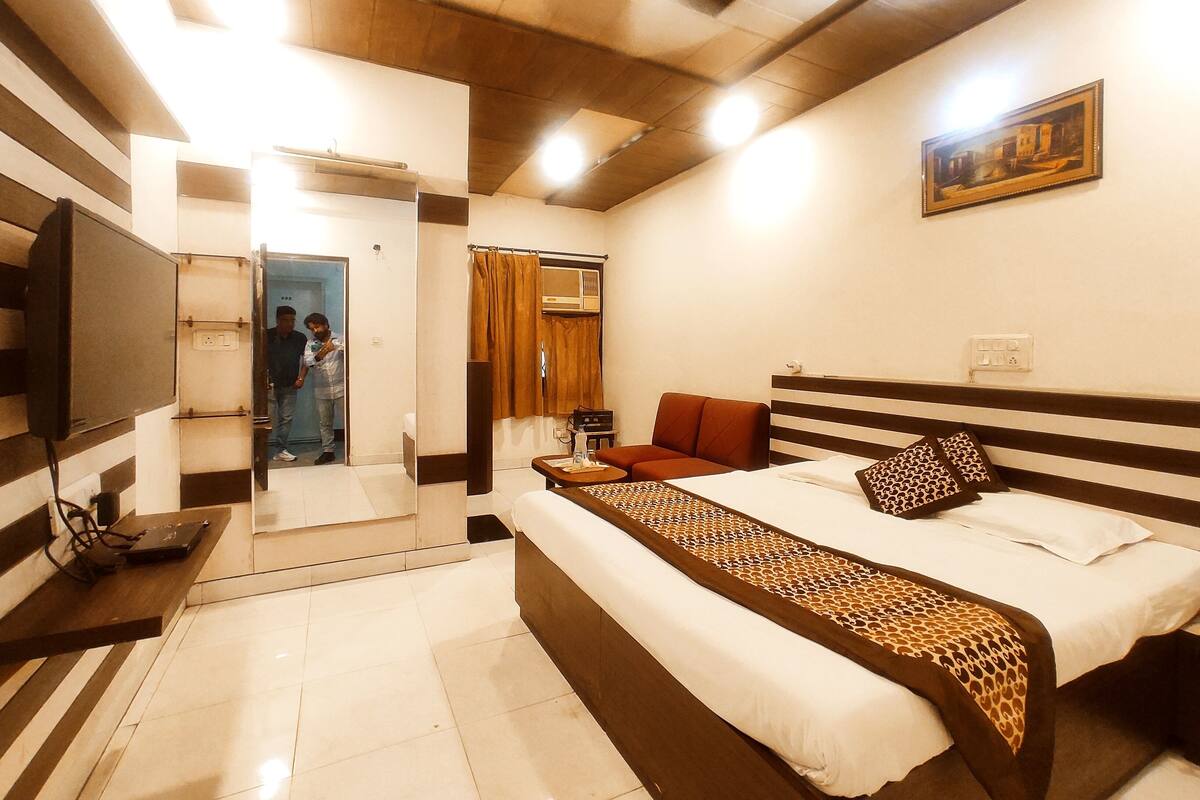 A hotel room with a bed, a sofa, a TV, a painting, and a bathroom visible through an open door.