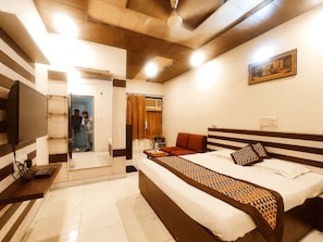 A hotel room with a bed, a sofa, a TV, a painting, and a bathroom visible through an open door. - THE SURYA PRIME Near AC Market (Ludhiana)