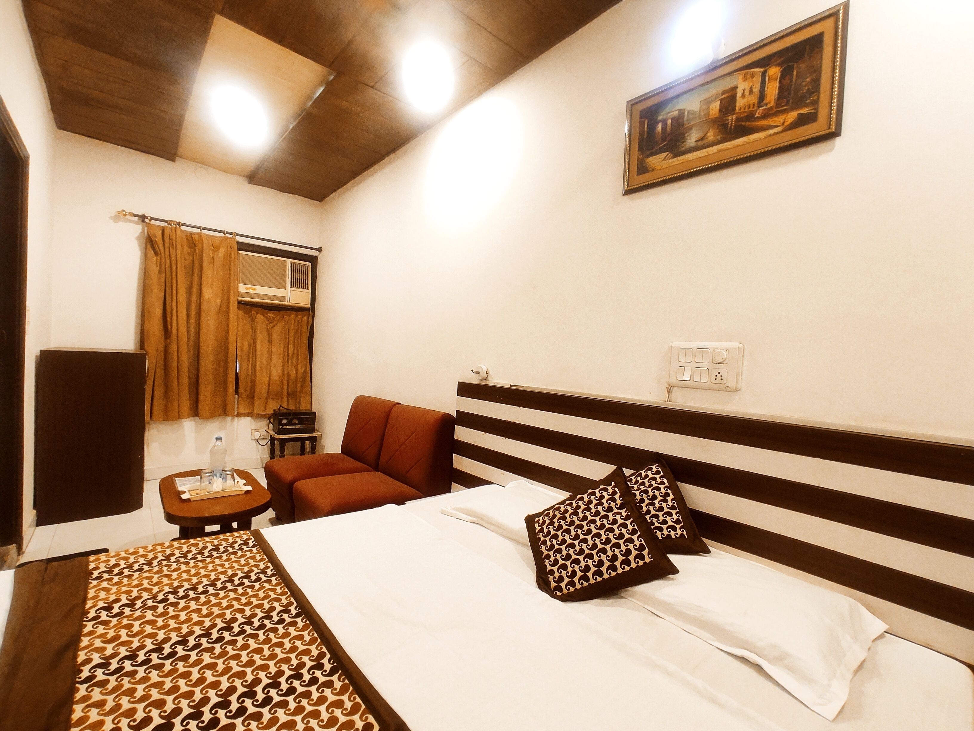Una habitación de hotel con cama, una silla, una mesa, un cuadro en la pared y un equipo de aire acondicionado.