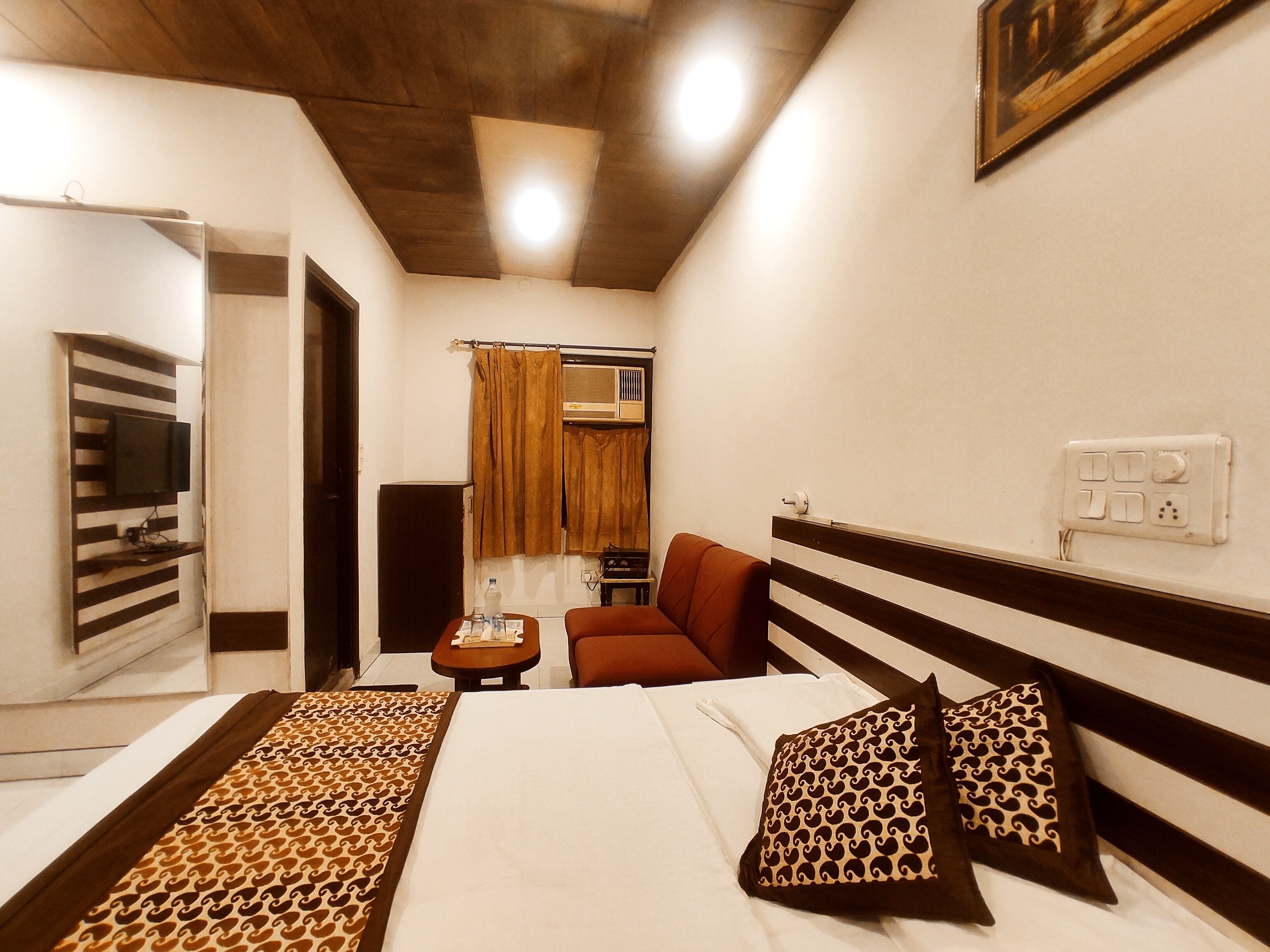 Habitación de hotel con cama, televisión montada en la pared, una silla, una mesita y una ventana con cortinas.