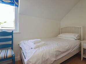 Una cama individual con sábanas blancas, una silla azul a rayas y una ventana con cortina azul.