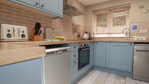 A kitchen with light blue cabinets, a wooden countertop, a built-in oven, a dishwasher, and a window with blinds. - Stowaway Cottage (Padstow)