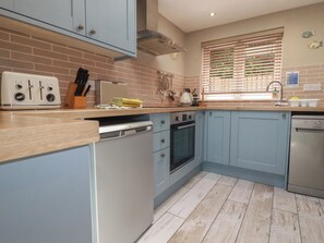 Une cuisine avec des meubles de couleur bleu clair, un plan de travail en bois, un four encastré, un lave-vaisselle et une fenêtre avec des stores.
