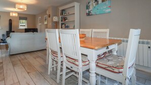 A dining area with a wooden table and white chairs, a white sofa, a bookshelf, and a painting on the wall. - Stowaway Cottage (Padstow)
