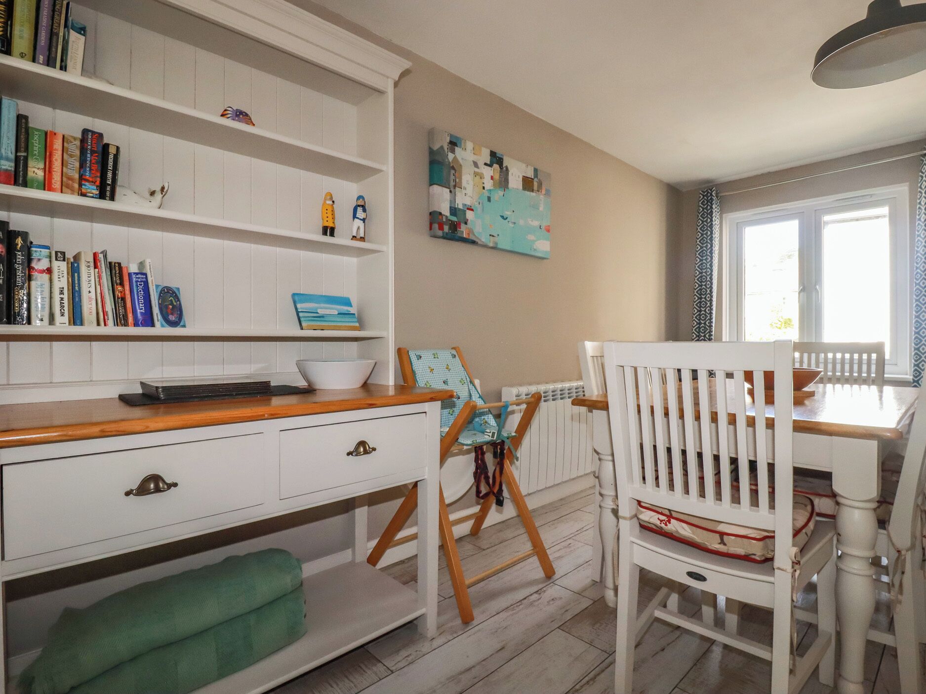 Una habitación con una estantería blanca, una mesa de comedor con sillas, un radiador y una ventana.