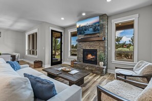 A living room with a fireplace, a sofa, a coffee table, and a view through large windows. - Brasada Ranchview Hot Tub | Fire Pit | Bunk Room (Powell Butte)