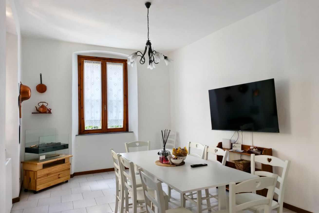 Zona pranzo con tavolo bianco, sedie, un lampadario, televisore a parete e una finestra con tende.