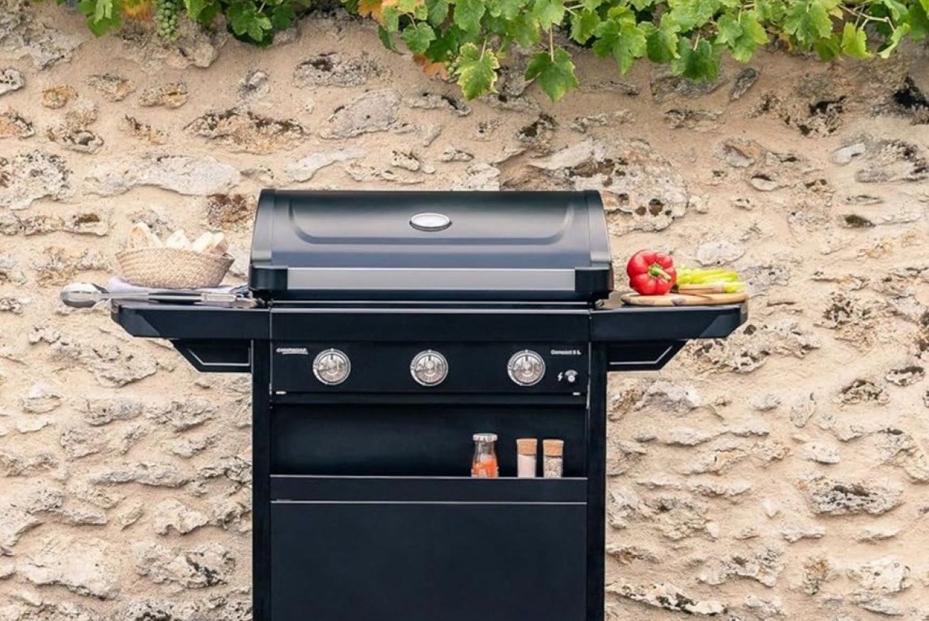 A black barbecue grill with three control knobs, a basket of bread, and a basket of vegetables against a stone wall.
