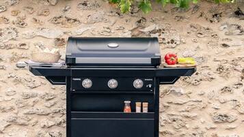A black barbecue grill with three control knobs, a basket of bread, and a basket of vegetables against a stone wall.