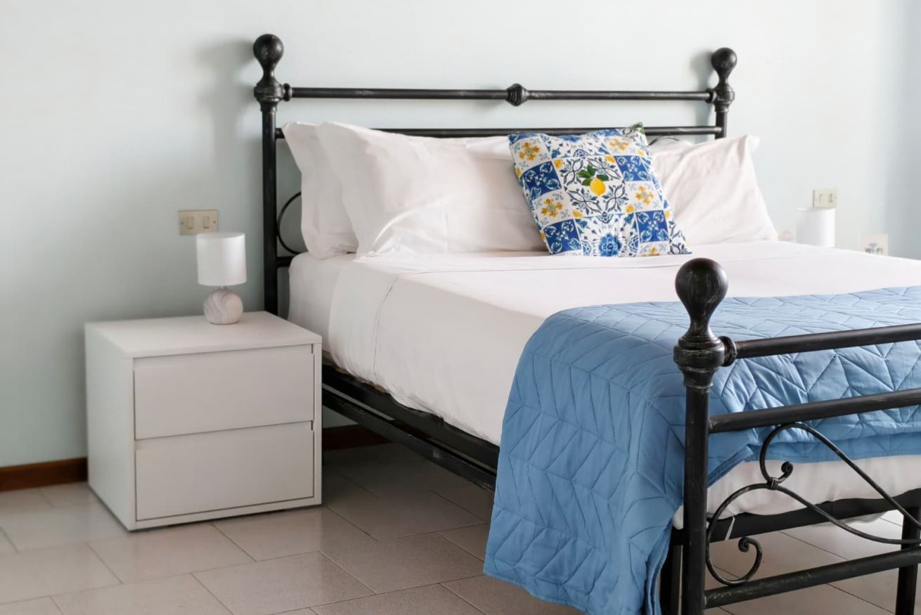 Un letto con struttura in metallo nero, biancheria da letto bianca e copripiumone trapuntato blu. È presente un comodino bianco con una lampada e un oggetto decorativo.