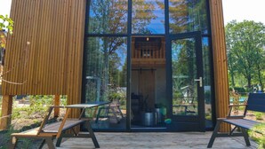 A modern wooden cabin with large glass windows, a deck with two chairs, and a visible interior with a hanging light fixture. - Holiday Home in Arnhem With Terrace (Arnhem)