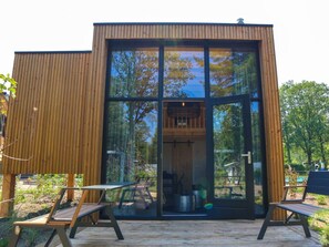 Eine moderne Holzhütte mit großen Glasfenstern, einer Terrasse mit zwei Stühlen und einem sichtbaren Innenraum mit einer Pendelleuchte.