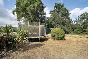 A trampoline enclosed by a safety net, surrounded by dry grass and various trees.