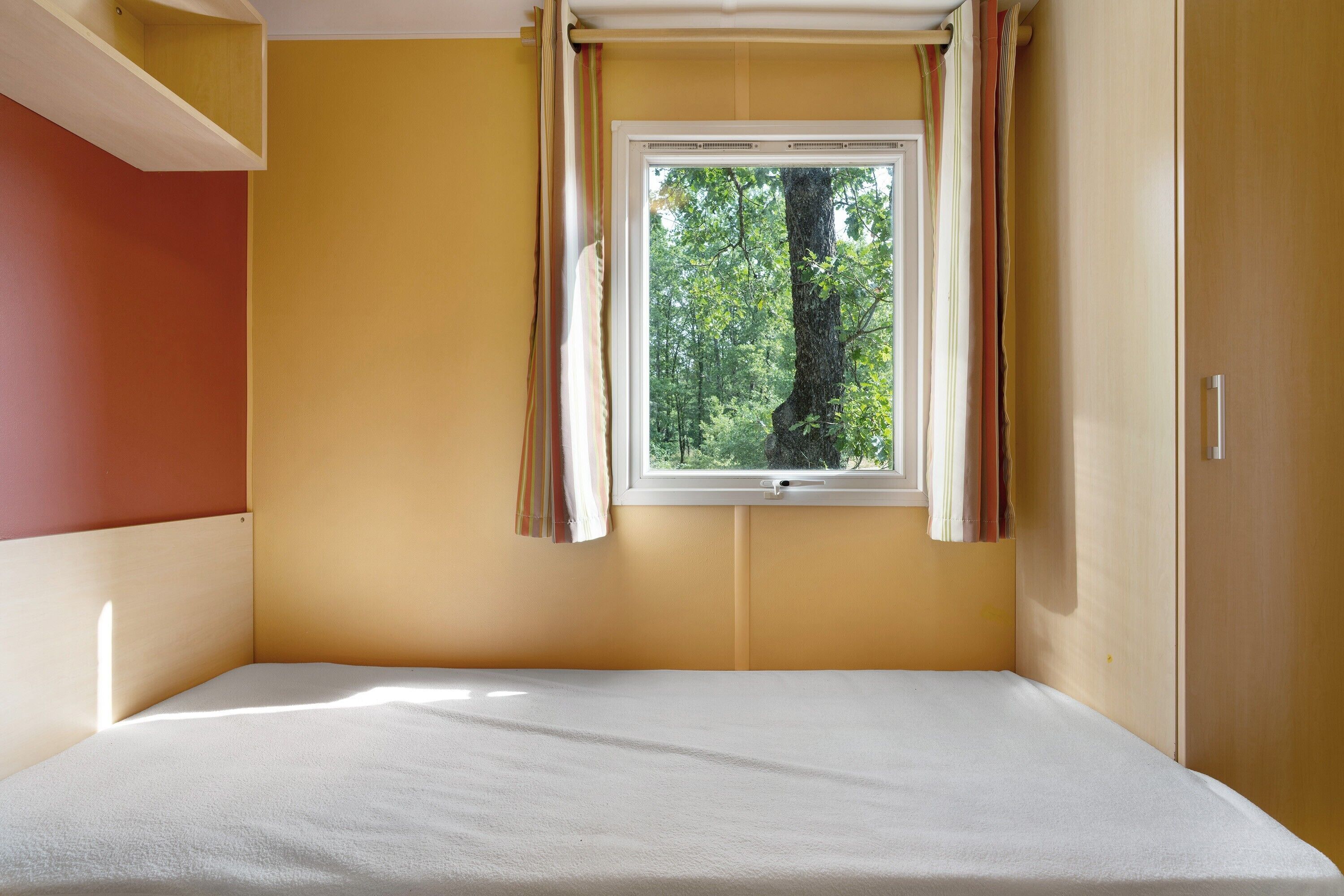 Una habitación con cama, ventana con cortinas y vista a árboles.