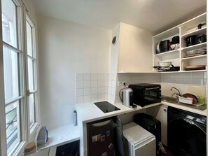 A compact kitchen with a microwave, toaster, and dishwasher, a small refrigerator, and a window with blinds. - Studio Jules Joffrin, dernier étage - Montmartre (Paris)
