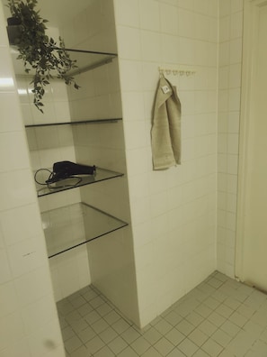 Kylpyhuone, jonka seinät on laatoitettu valkoisilla laatoilla, ja jossa on lasihylly, seinälle ripustettu pyyhe ja ruukussa oleva kasvi.