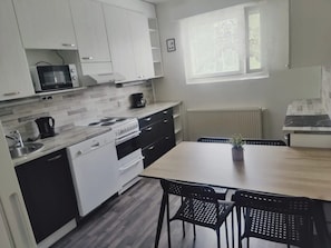 Kompakti keittiö, jossa on valkoiset kaapit, mikroaaltouuni, liesi ja ruokapöytä tuoleineen.