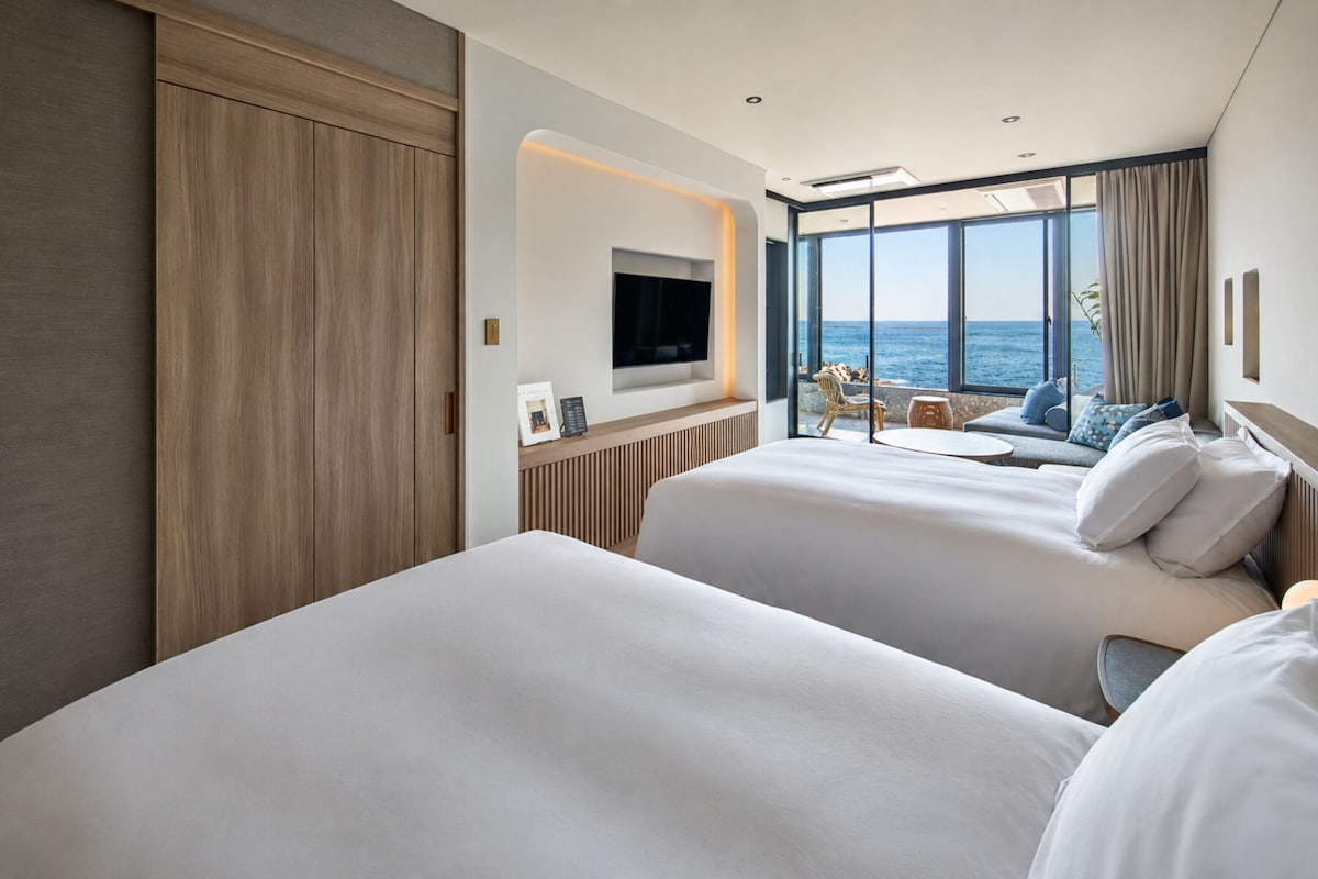 A hotel room with two beds, a TV, and a view of the sea.
