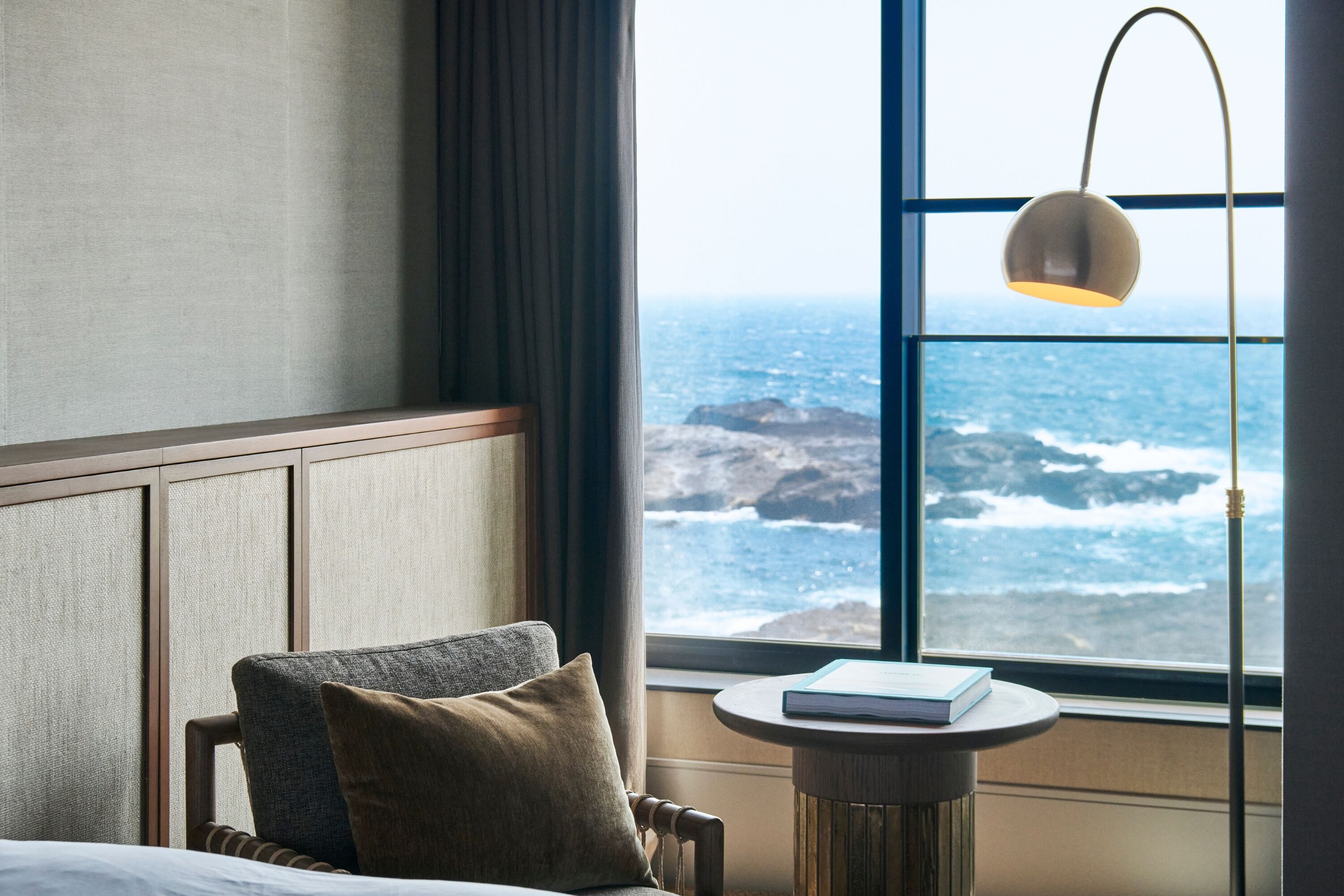 A hotel room with a bed, a chair, a small table with a book, a window with a view of the ocean, and a hanging lamp.