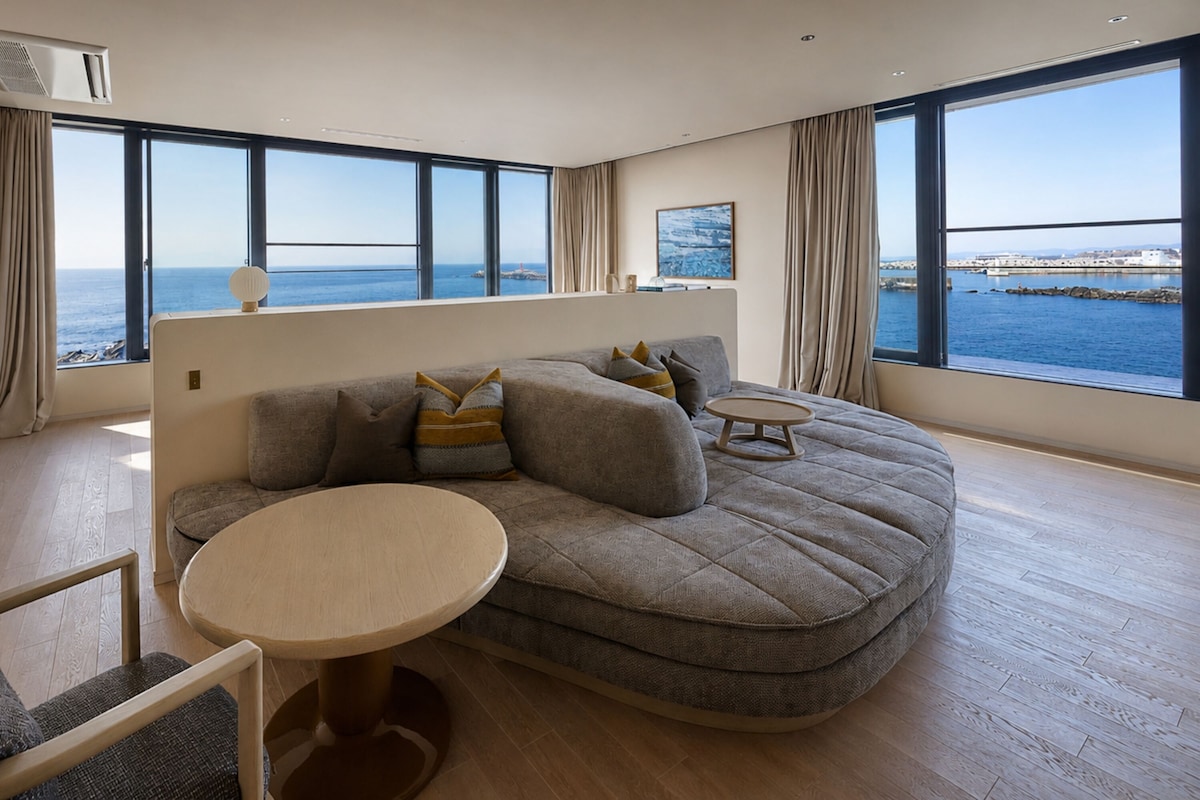 A modern living room with a large window, a sofa, a round coffee table, and a view of the sea.