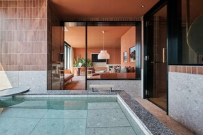 Un área de piscina moderna al aire libre con cerramiento de vidrio, una zona de estar con un sofá y una mesa de comedor, y una cocina visible a través del vidrio.