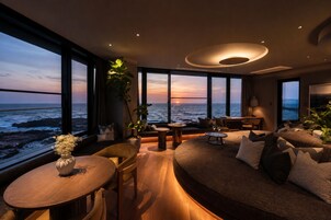 A modern living room with a view of the ocean at sunset, featuring a sofa, a coffee table, and a dining area.