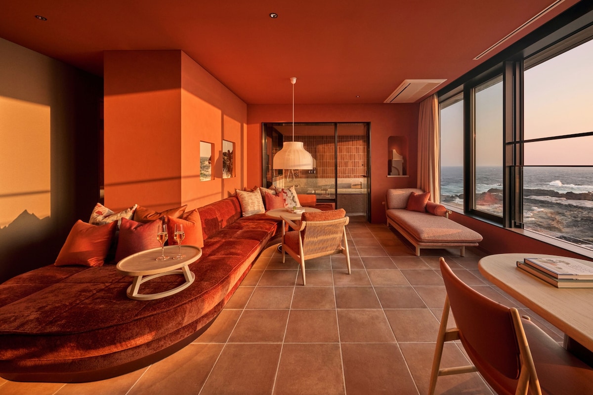 A modern living room with a large window, a sofa set, a dining area, and a view of the sea.