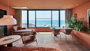 Una habitación de hotel moderna con un ventanal grande con vista al océano, un sofá, una mesa de comedor con sillas y una planta pequeña.