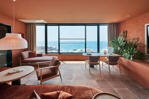 A modern hotel room with a large window overlooking the ocean, a sofa, a dining table with chairs, and a small plant.