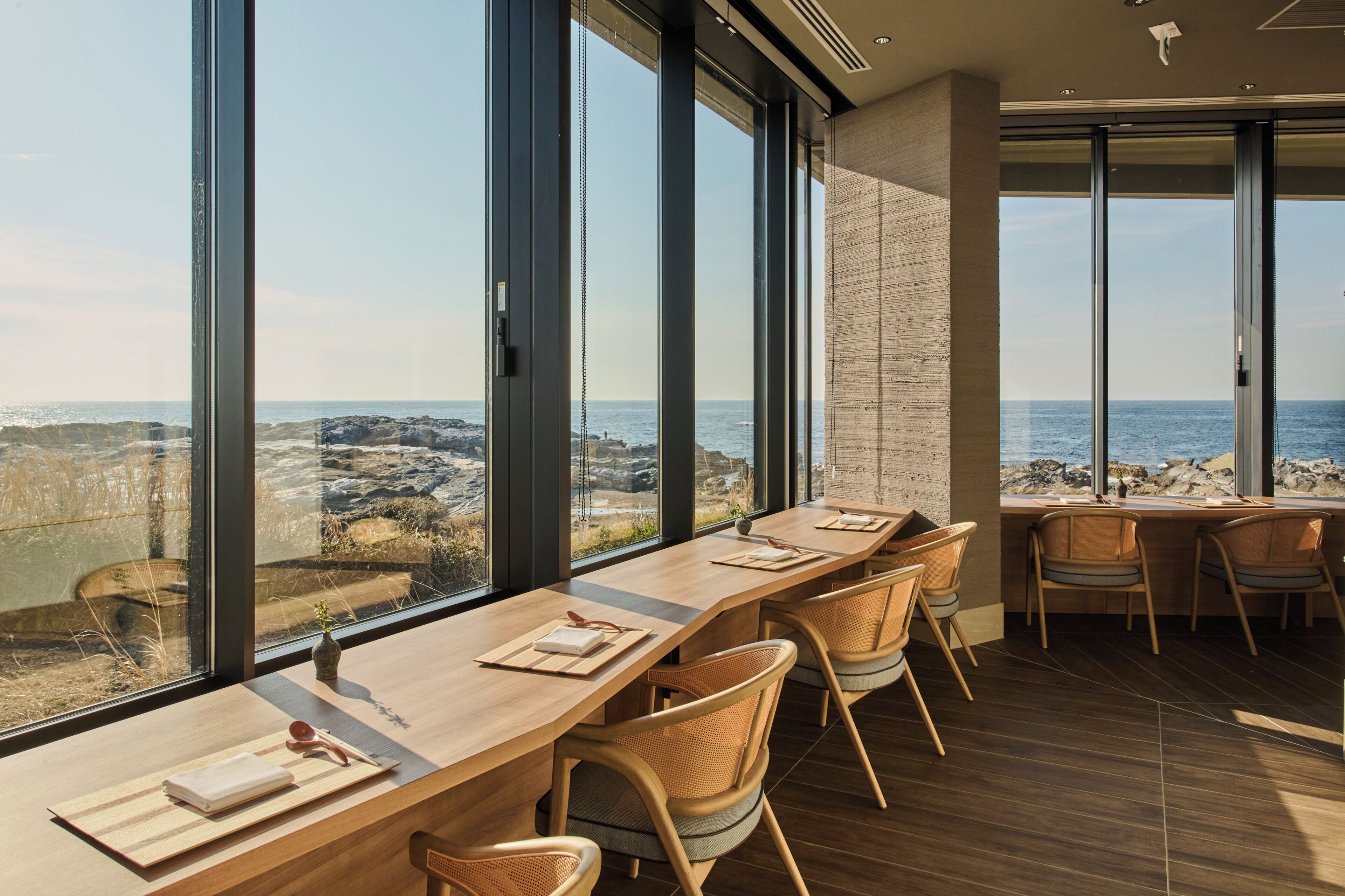 A modern conference room with a long table, chairs, and large windows offering a view of the ocean and rocky landscape.