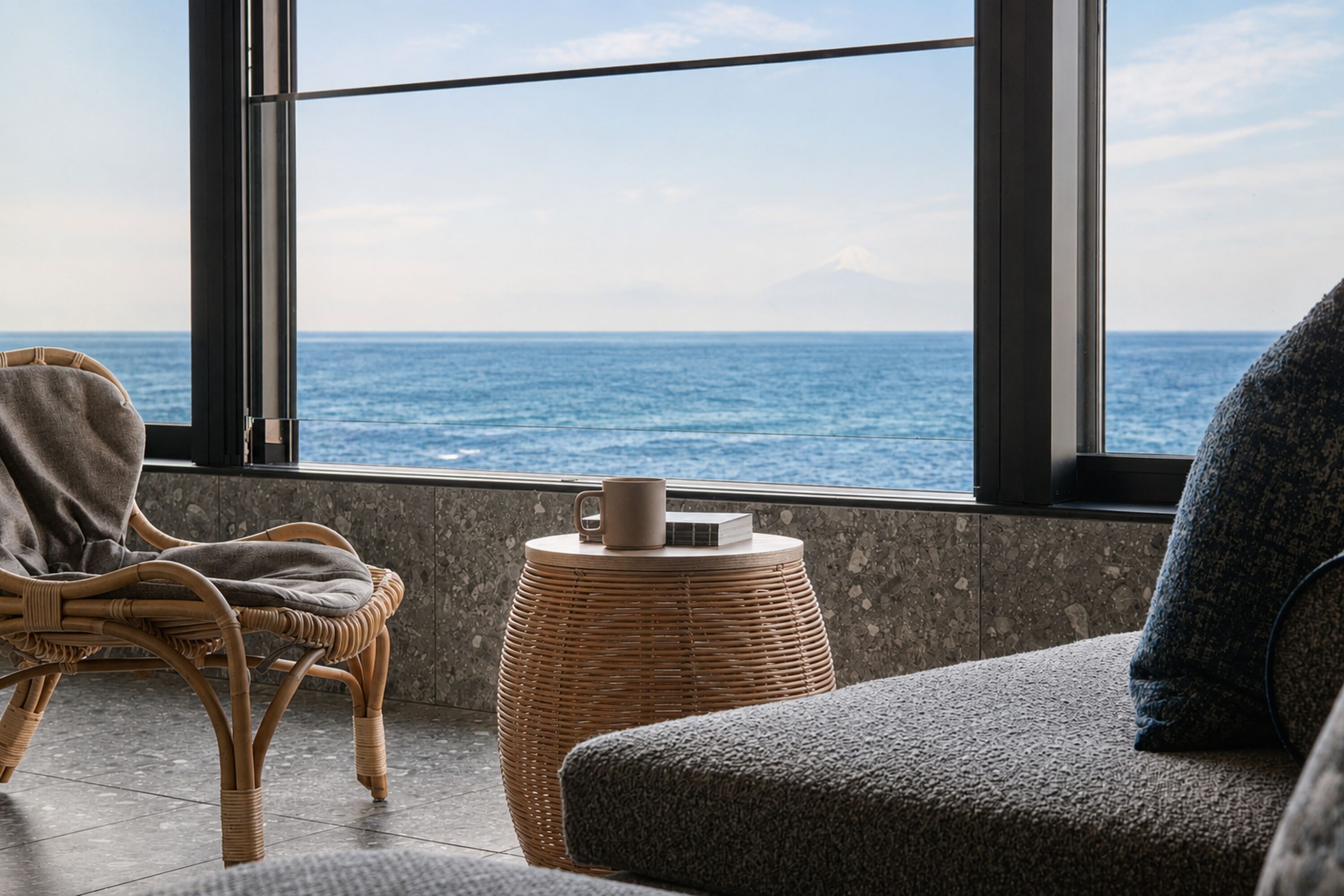 A room with a view of the sea, a wicker chair, a small round table with a mug, and a grey sofa.