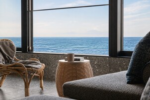 A room with a view of the sea, a wicker chair, a small round table with a mug, and a grey sofa.