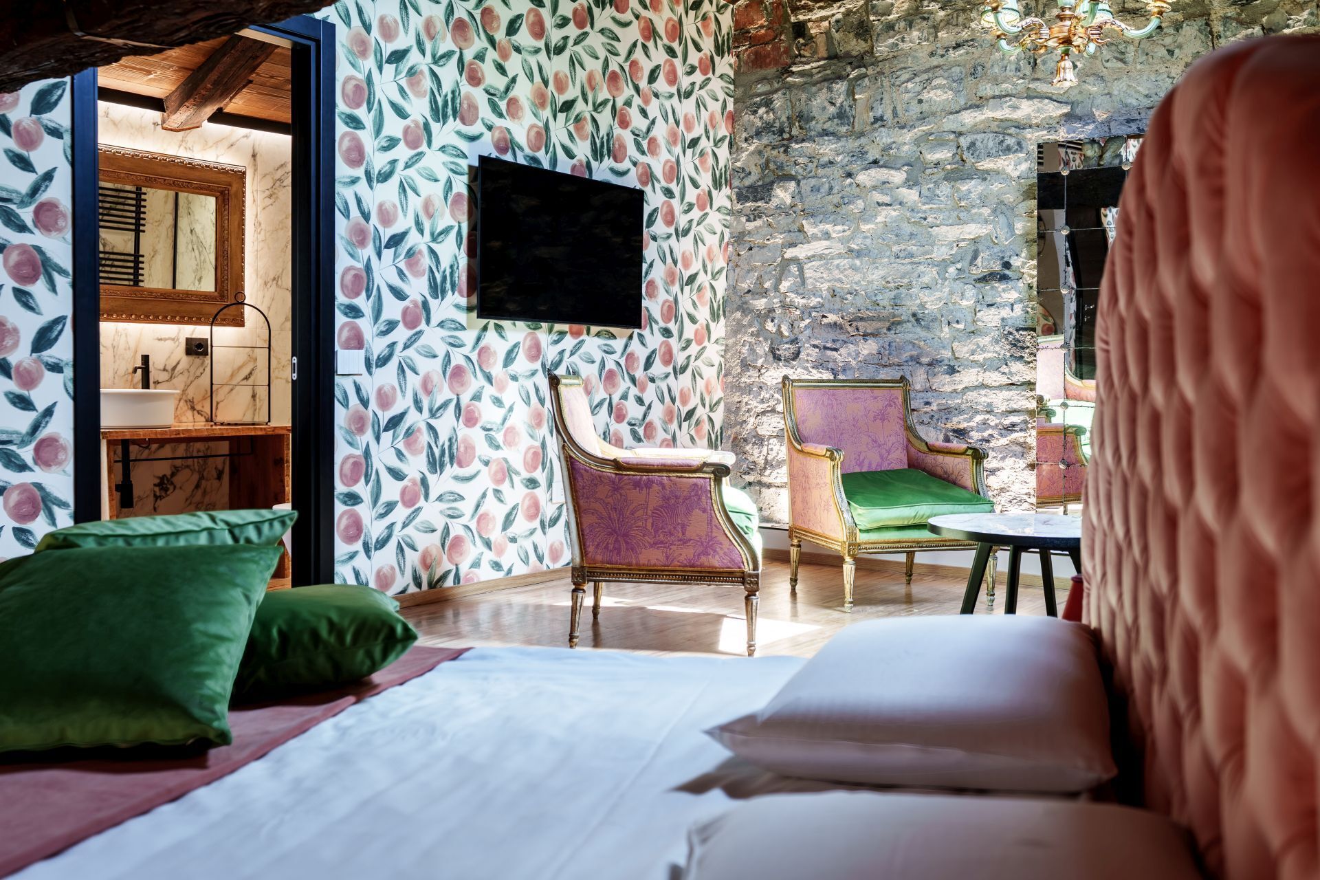 Una habitación con cama, dos sillas, una mesita, un espejo y una pared de piedra.