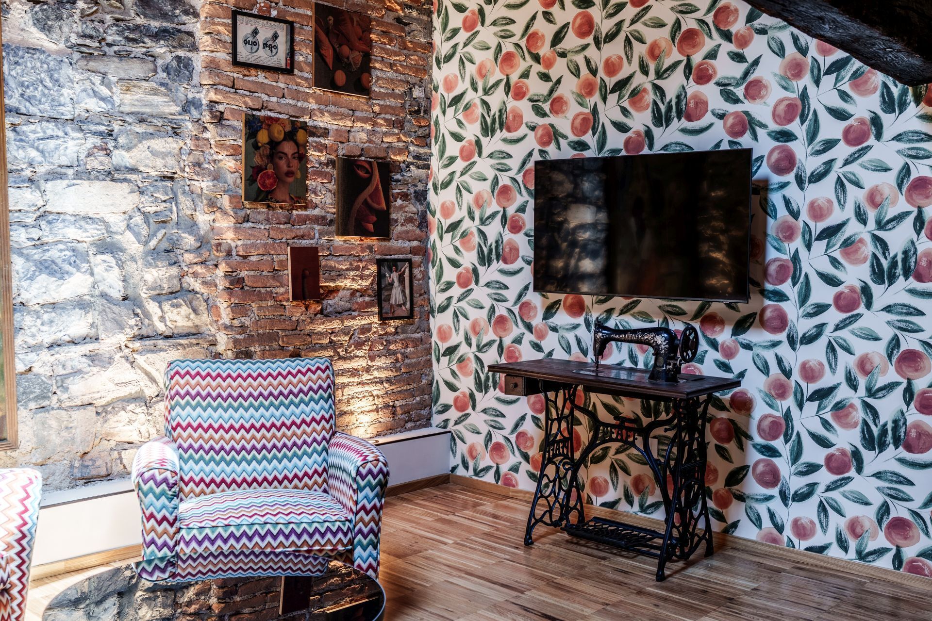 A room with a brick wall, a patterned wallpaper, a television, a sewing machine, and a colorful chair.