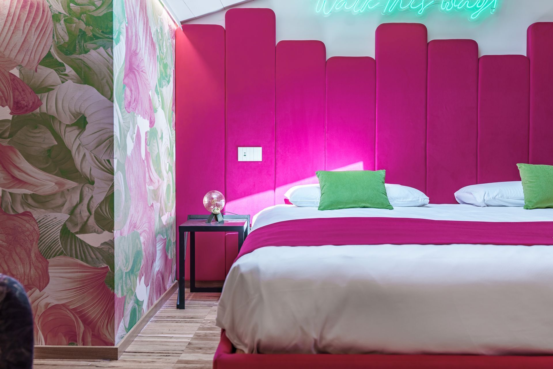 A modern hotel room with a vibrant pink accent wall, a bed with white and green pillows, and a neon sign.