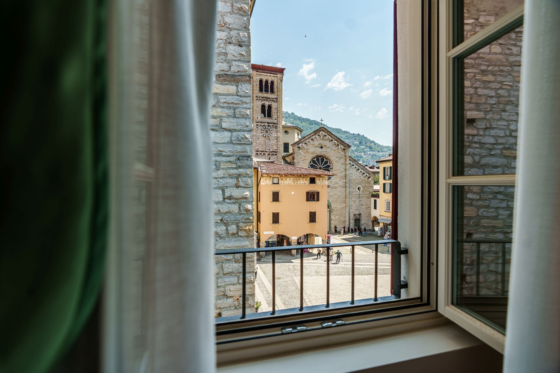 Blick durch ein Fenster mit Balkon, zeigt ein Steinhaus mit Glockenturm und Turuhr sowie einen gepflasterten Platz mit Menschen.