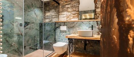 A modern bathroom with a glass shower enclosure, a freestanding bathtub, a sink with a wooden countertop, and a stone wall backdrop.