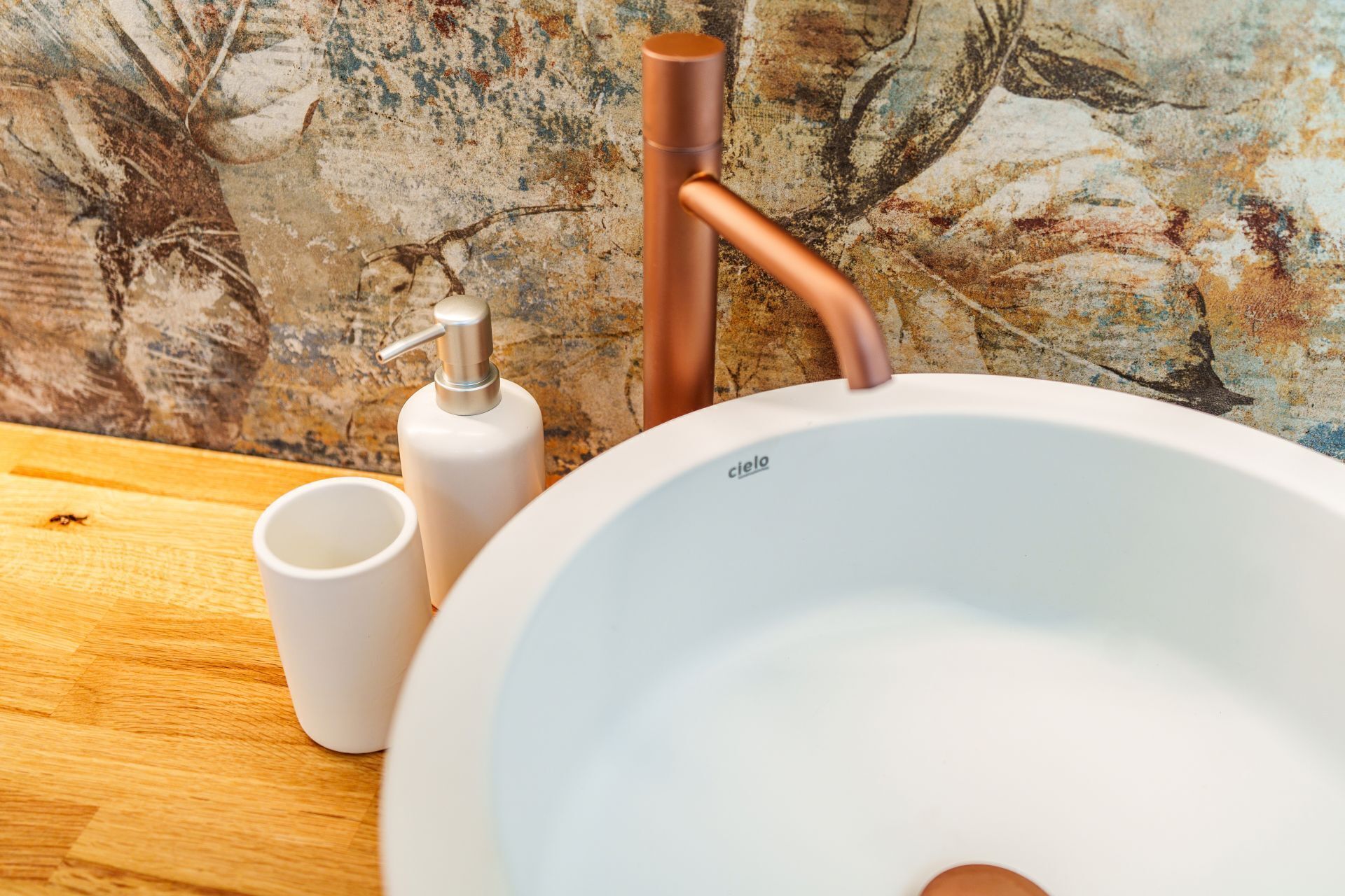 A bathroom sink with a copper faucet, a white basin, and a white cup on a wooden surface.